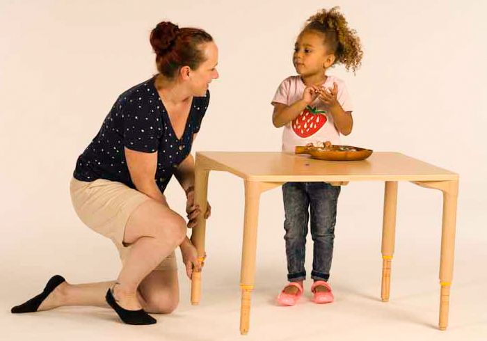 Ein kleines Kind steht an einem Tisch, während eine Erzieherin die Tischhöhe verstellt