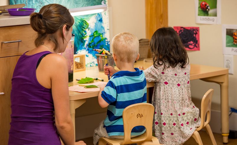 Eine Erzieherin schaut auf zwei Kinder, die mit Buntstiften und Kunstmaterialien an einem Tisch sitzen.