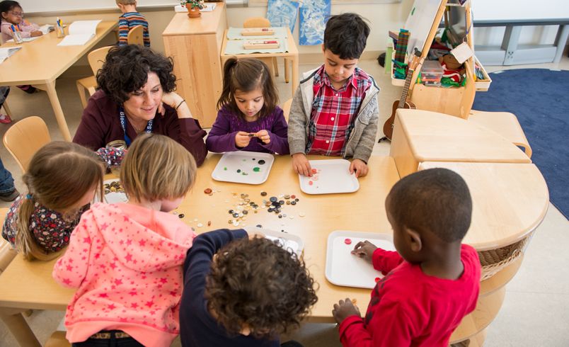 Eine Gruppe von Kindern und eine Erzieherin sitzen oder stehen um einen Tisch herum und spielen mit farbigen Glasperlen.